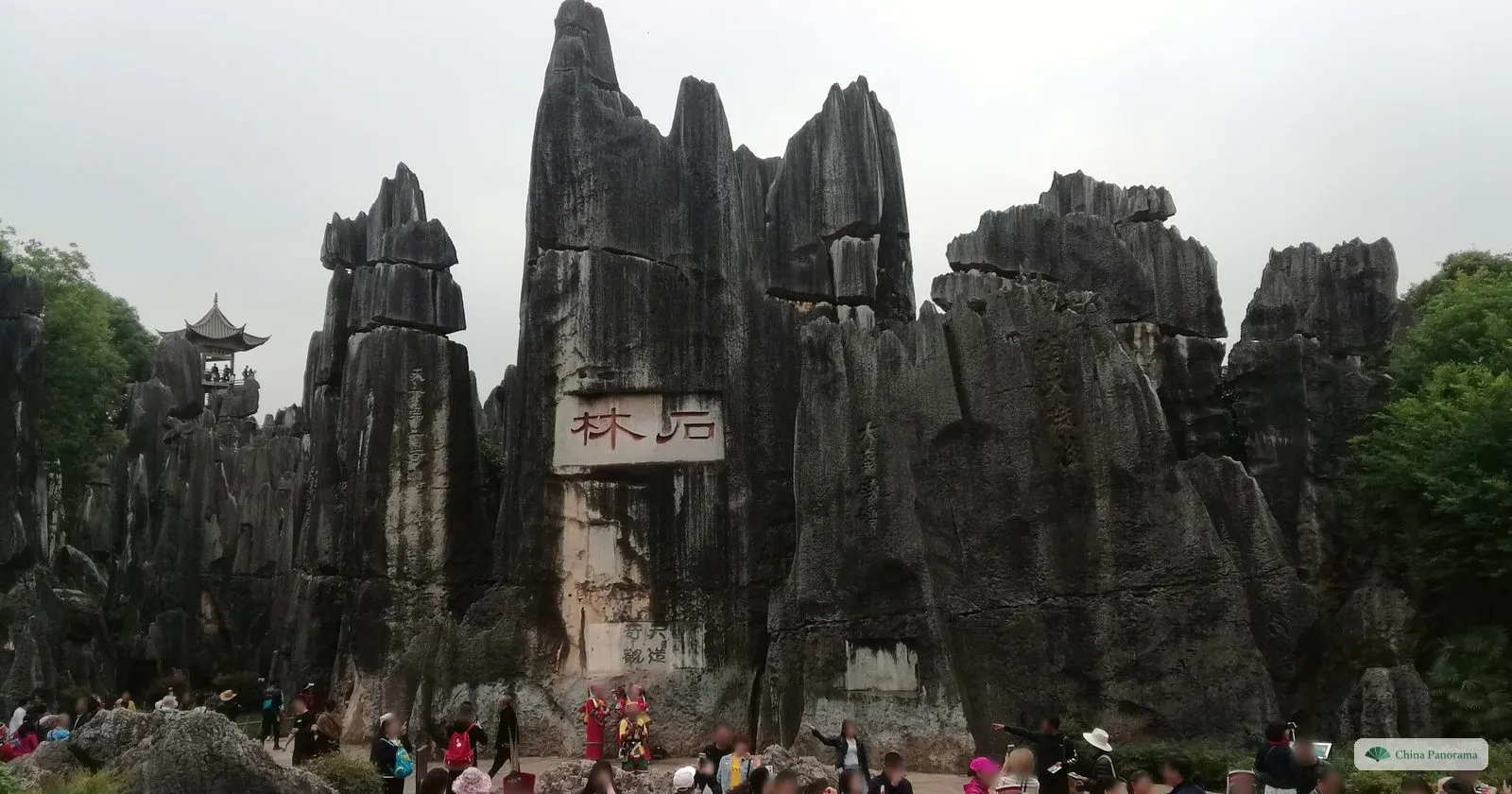Tall dark rock pinnacles under a grey sky, one carved with two large red characters on a white panel, a small pavilion on a rock top to the left and visitors below