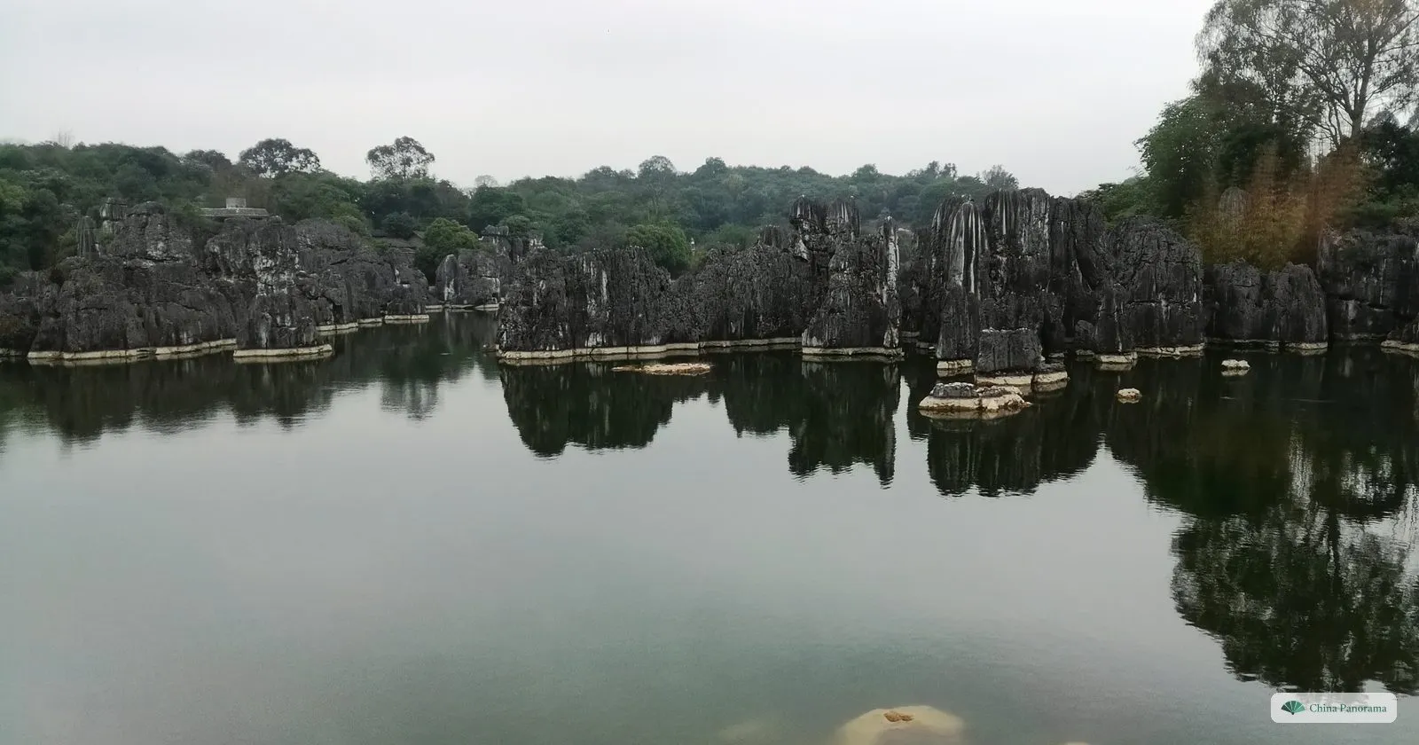 A long line of low grey rocks along the far shore of a calm lake, mirrored in the water, a cloudy sky