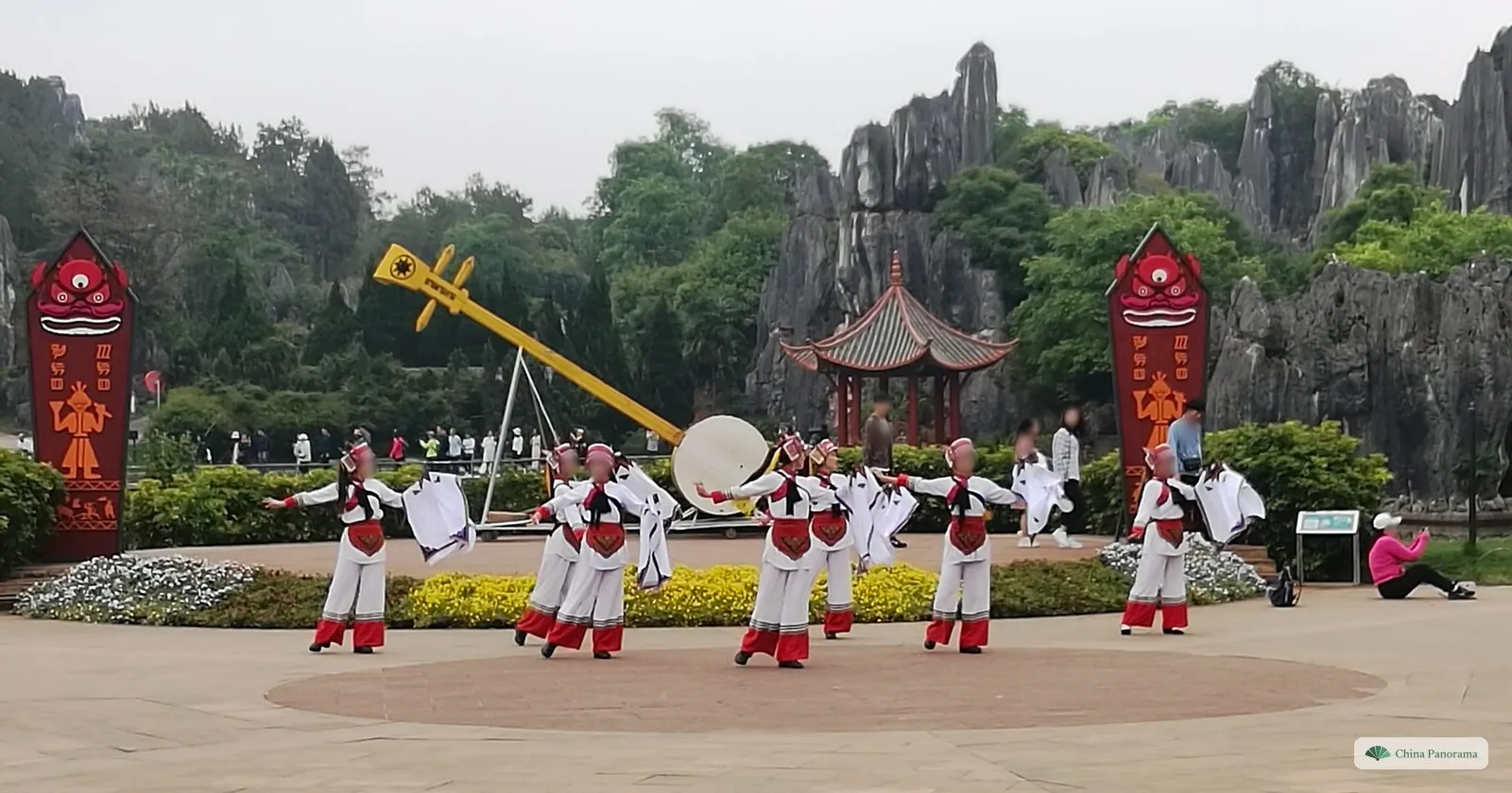 Dancers in white, red and black costumes waving white cloths on a paved square beside a giant model of a three-stringed lute and a drum, rock pinnacles behind