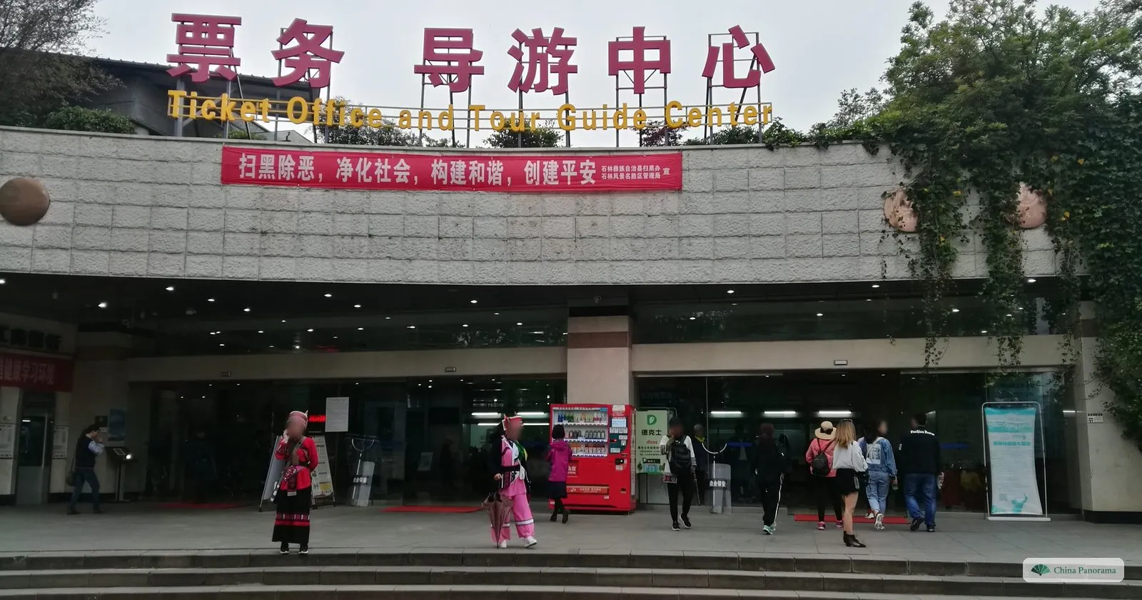 The entrance of a visitor centre with large red characters and the words Ticket Office and Tour Guide Center, women in colourful costume at the door