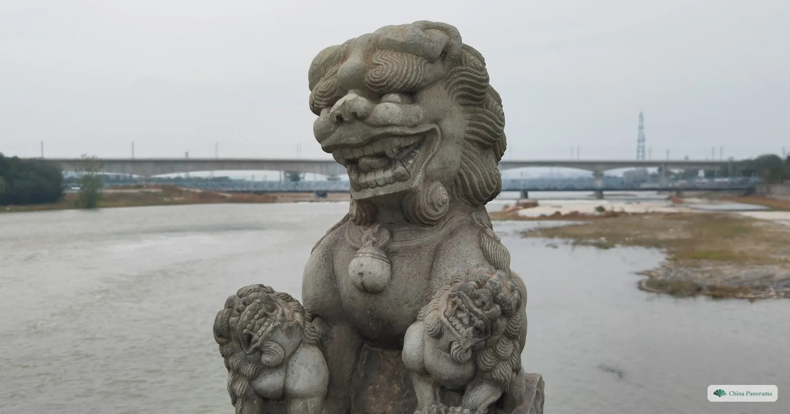 A stone lion with two cubs on the parapet of Lugou Bridge, with the Yongding River and a modern bridge behind