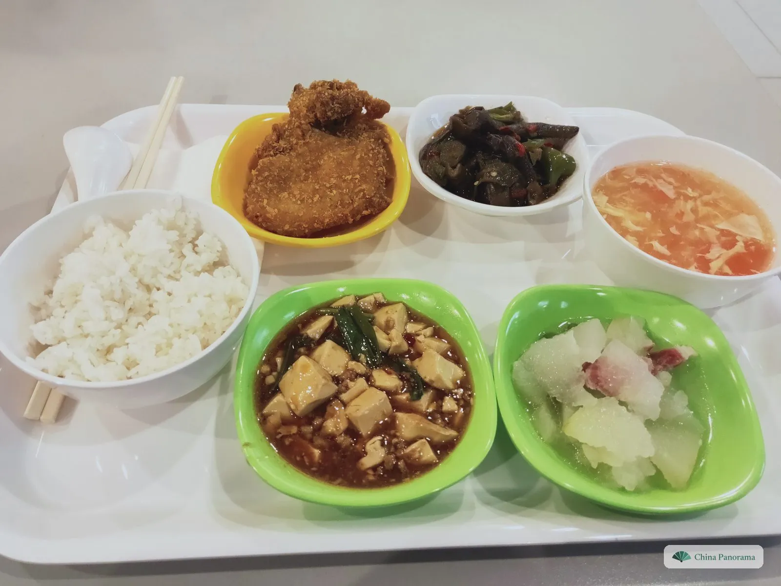 A canteen tray from above: a bowl of rice, a breaded cutlet in a yellow bowl, eggplant with green pepper in a small white bowl, tomato and egg soup, and two green bowls – tofu in red sauce, and winter melon