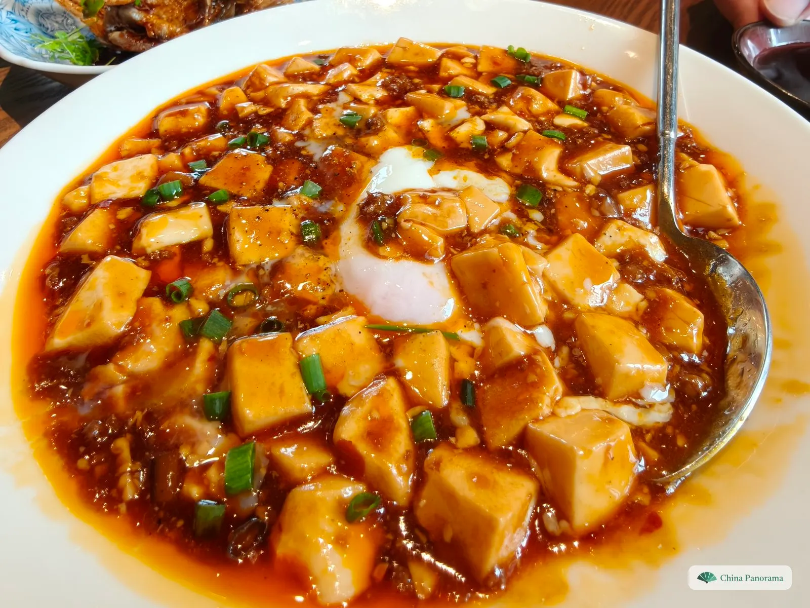 Close-up of a large plate of mapo tofu: soft tofu cubes in a glossy red chili sauce with minced meat and chopped spring onion, a soft-cooked egg in the middle and a metal spoon on the right