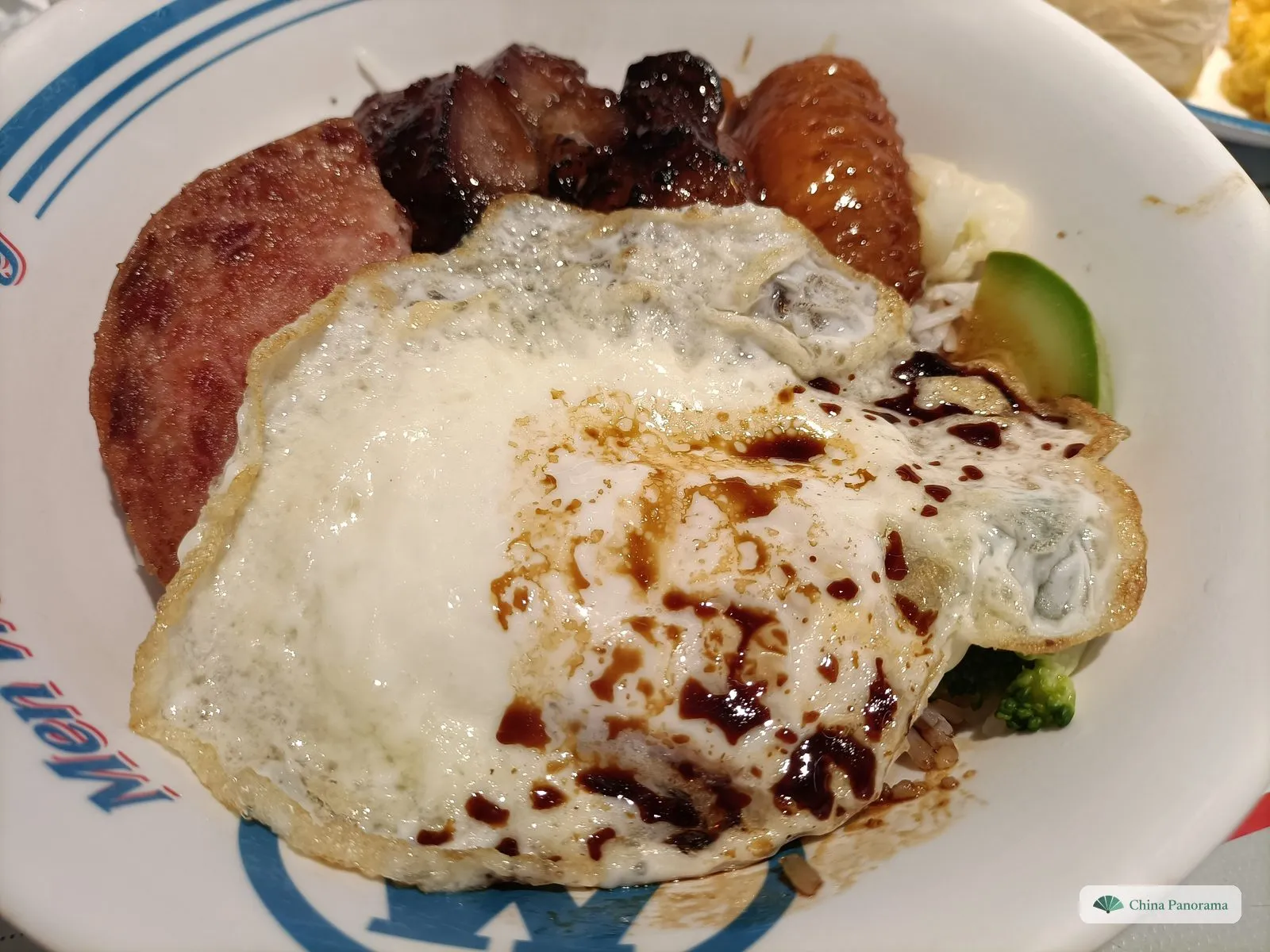 A bowl of rice topped with roast meats – dark char siu at the back – and a fried egg drizzled with sweet soy sauce