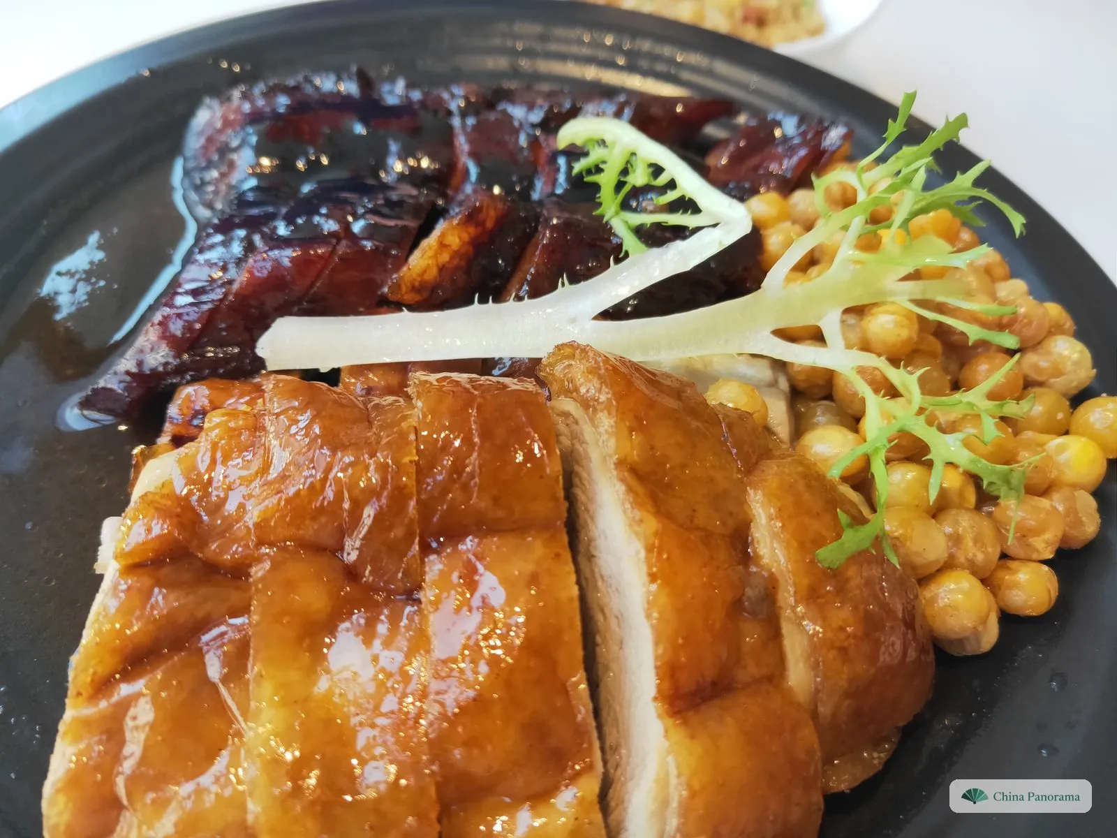 A black plate with two roast meats: dark, lacquered strips of pork at the back and golden slices of roast goose with crisp skin in front, with chickpeas and frisée on the right