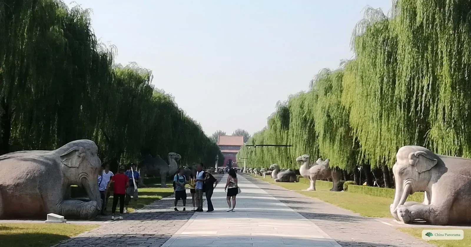 Two kneeling stone elephants on either side of the Sacred Way of the Ming Tombs, with camels behind and a few visitors on the path