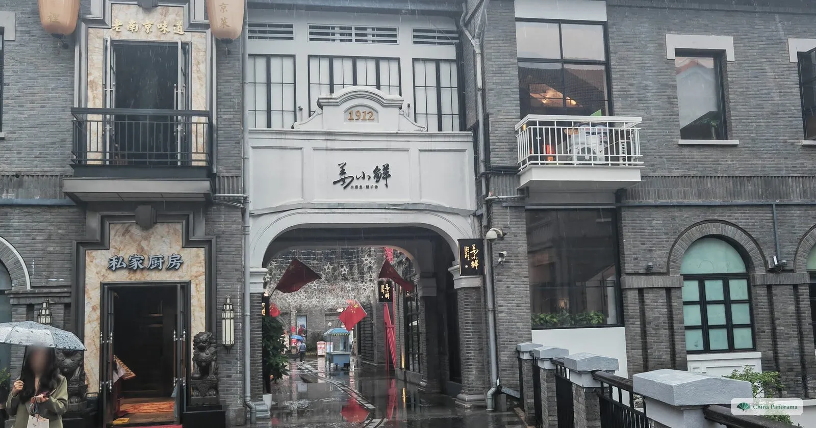 A white gatehouse marked 1912 between grey brick buildings, a passage hung with red flags and lights leading through it, wet pavement
