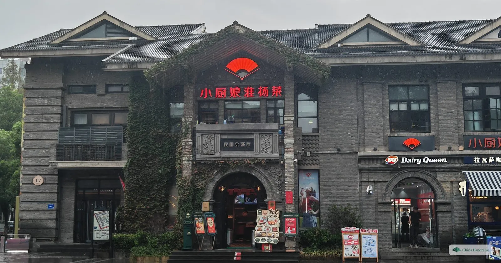 A grey brick building of the early twentieth century with arched windows, ivy and restaurant signs, a wet paved square in front