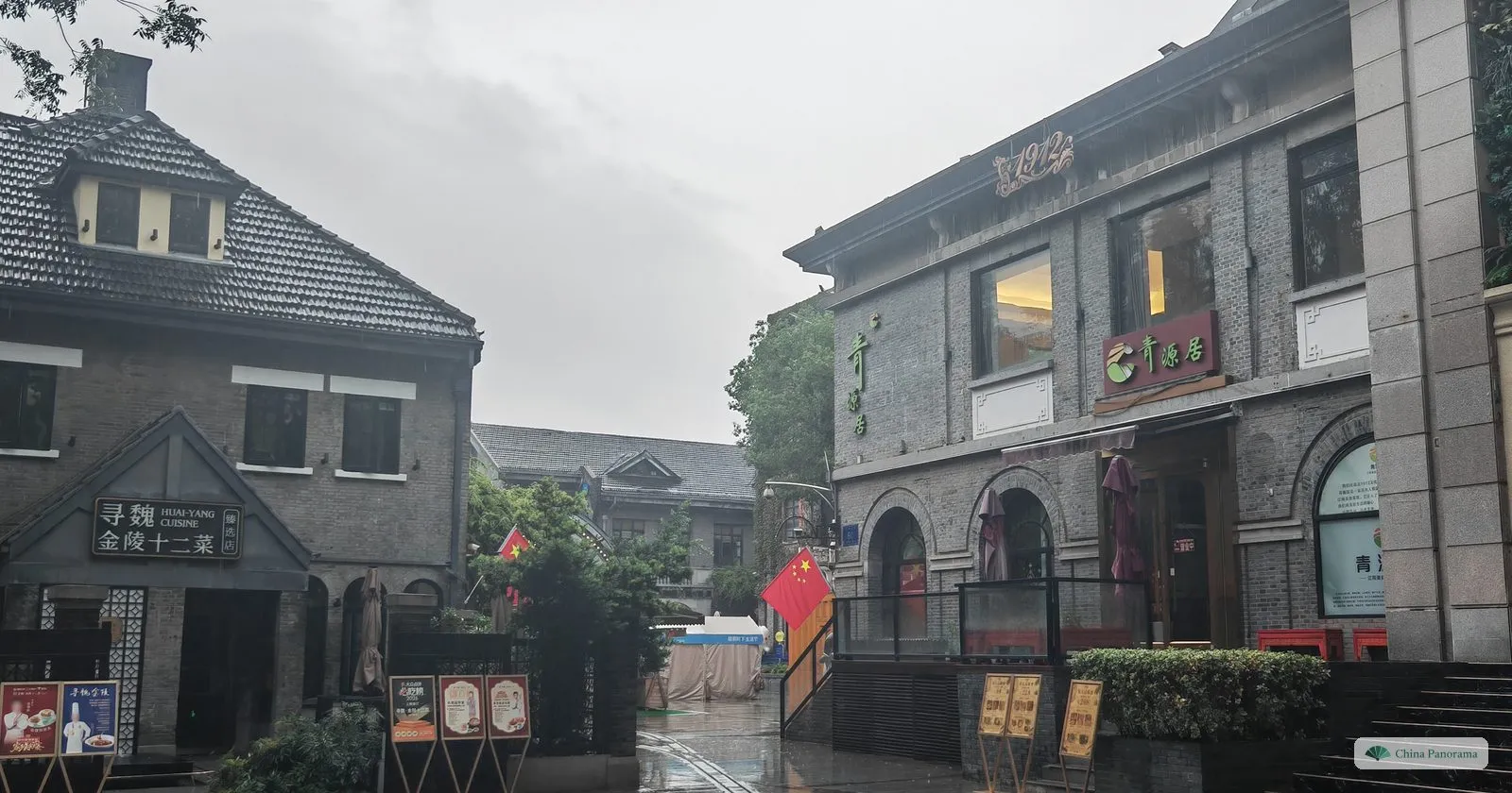 Grey brick houses with gabled roofs and arched windows along a wet lane, restaurant boards and a red flag