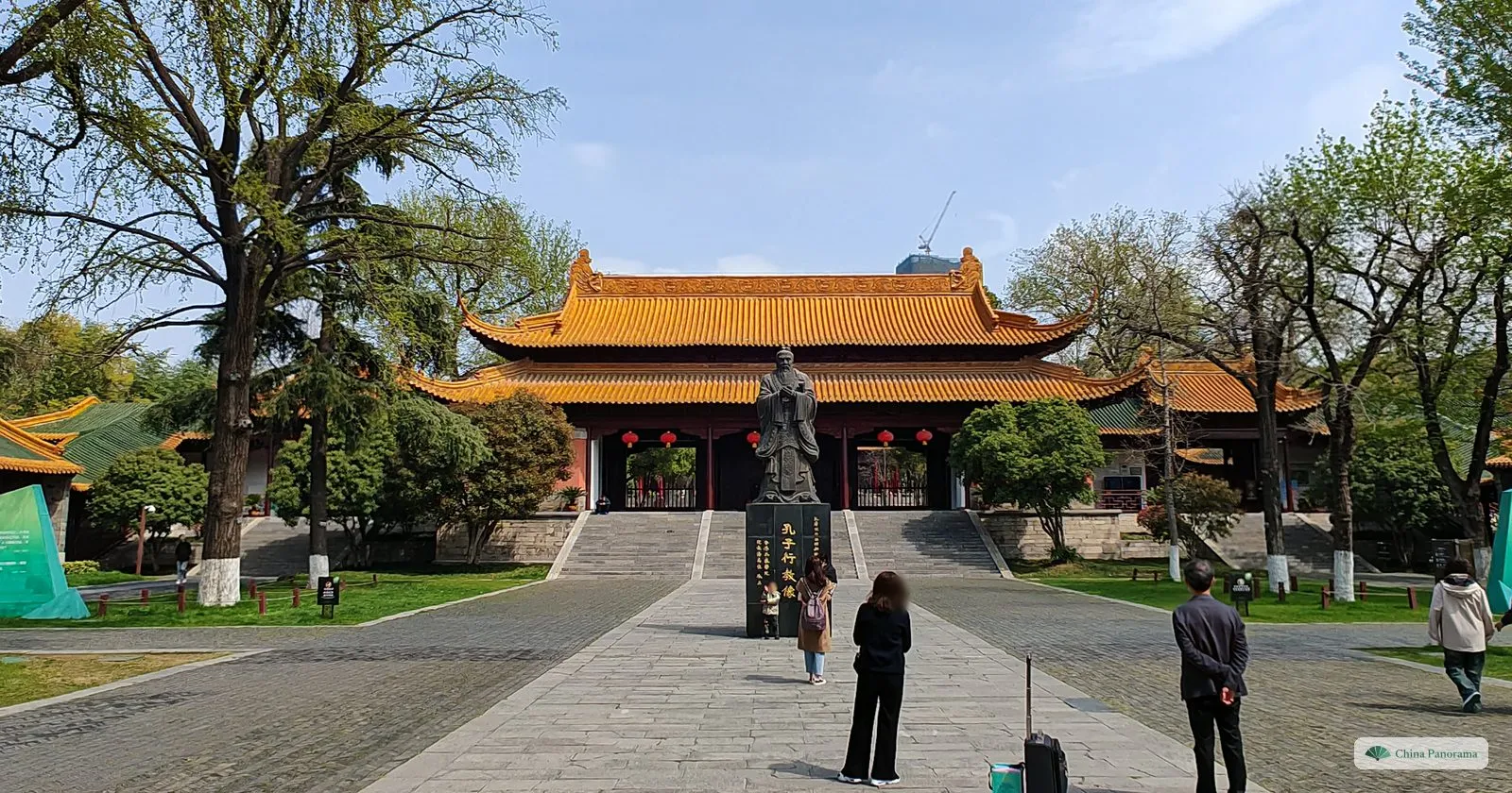 A bronze statue of Confucius on a black pedestal in front of a gate hall with yellow tiled roofs, visitors on the paved path