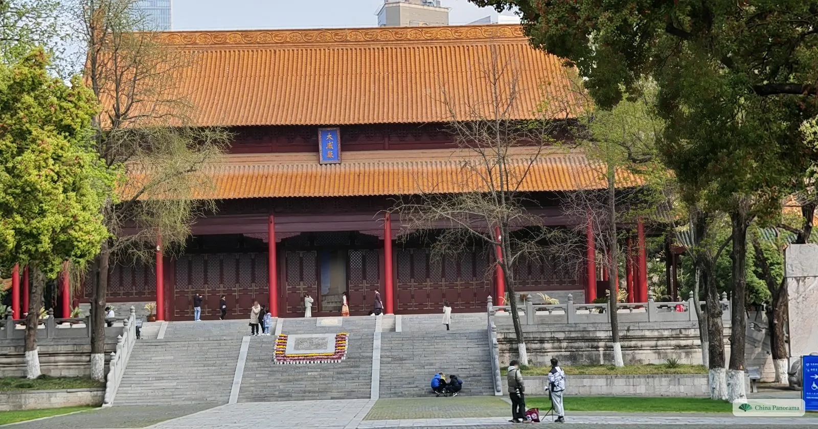 A large hall with a double roof of yellow glazed tiles and red columns on a stone terrace, a blue vertical plaque under the eaves and trees either side