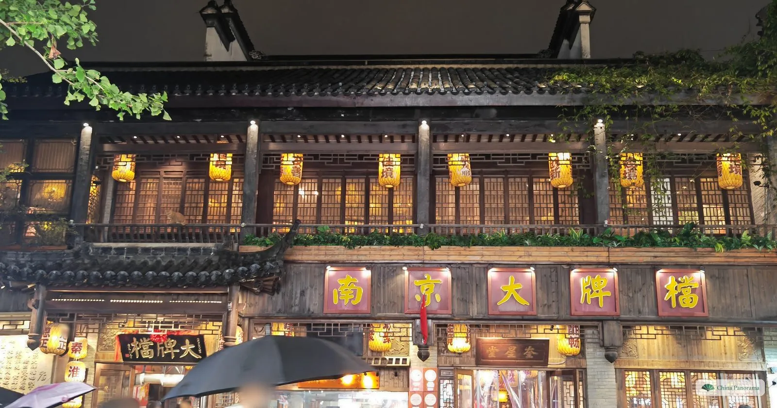 A two-storey wooden restaurant front at night with yellow lanterns on the balcony and red boards reading 南京大牌檔