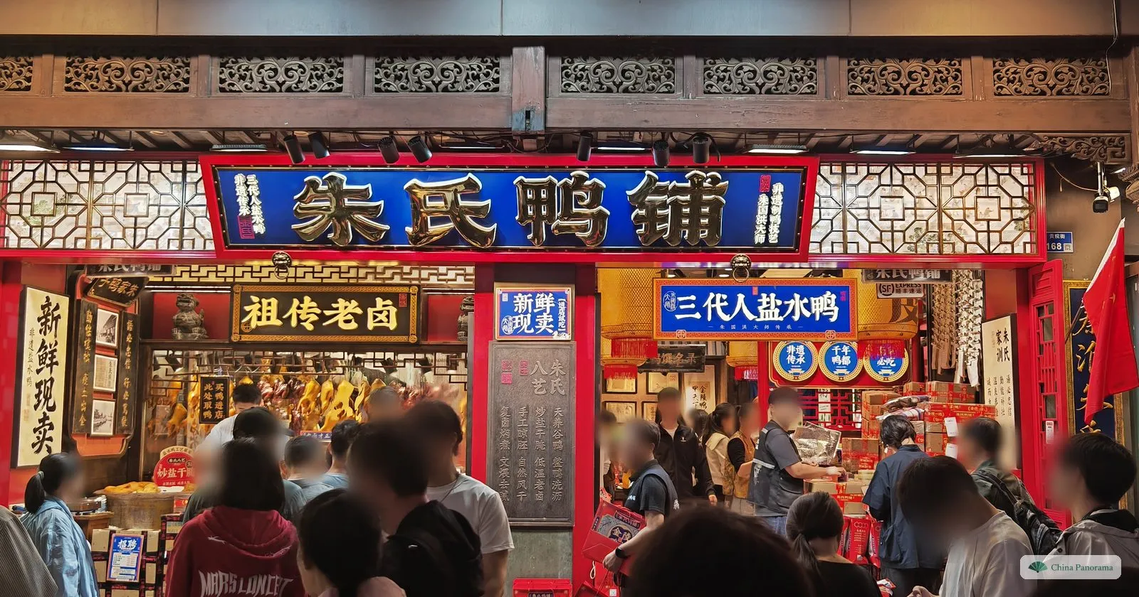 A shopfront at night with a blue sign 朱氏鸭铺 in gold characters, roast ducks hanging inside and a crowd of customers