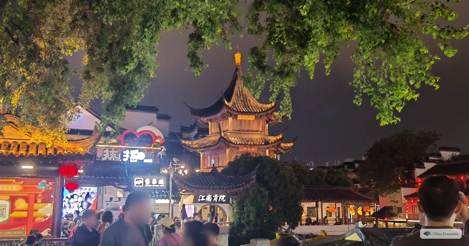 A three-storey octagonal pavilion lit gold at night above shop signs, with ginkgo branches overhead