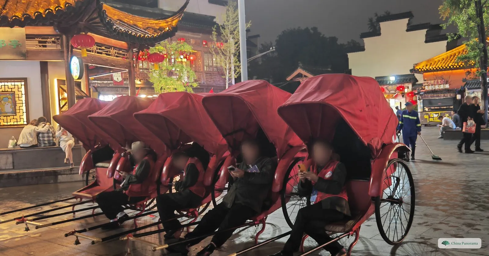 A row of red covered rickshaws parked on a wet square at night, their drivers resting in the seats