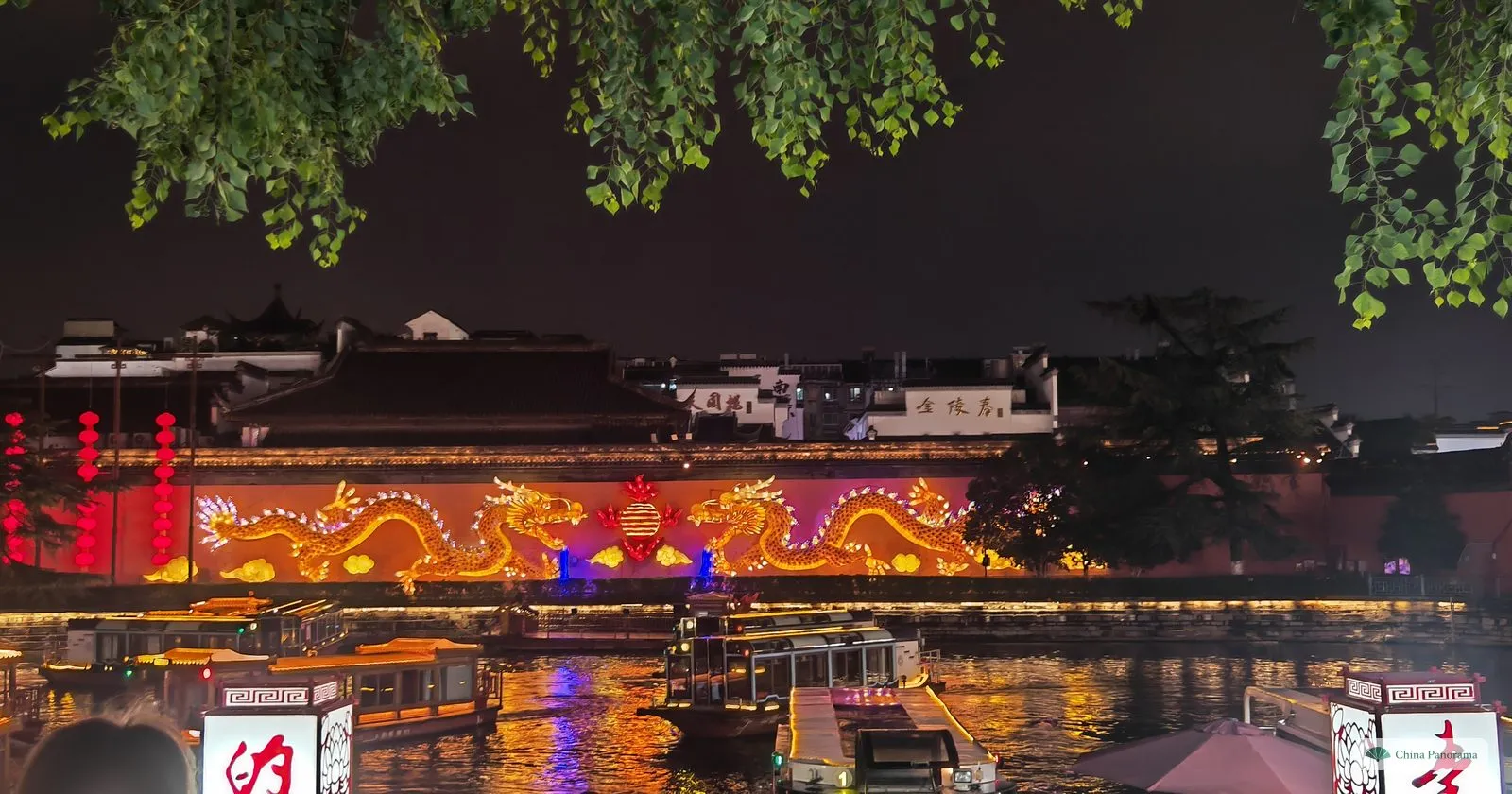 A long wall across a river at night, lit with golden dragons and red lights, reflections in the water and boats moored below