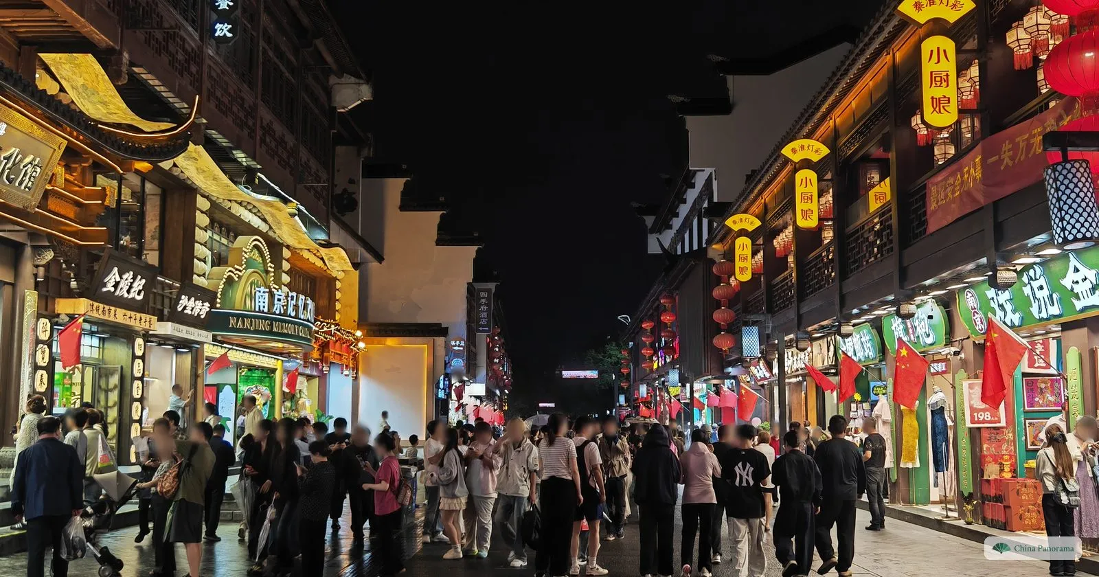 A crowded pedestrian street at night between brightly lit shops and restaurants with red lanterns and flags
