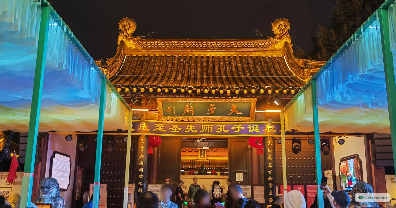 A temple gate lit gold at night under a canopy of blue light, a green plaque 夫子廟 and a banner below it, a crowd at the ticket gates