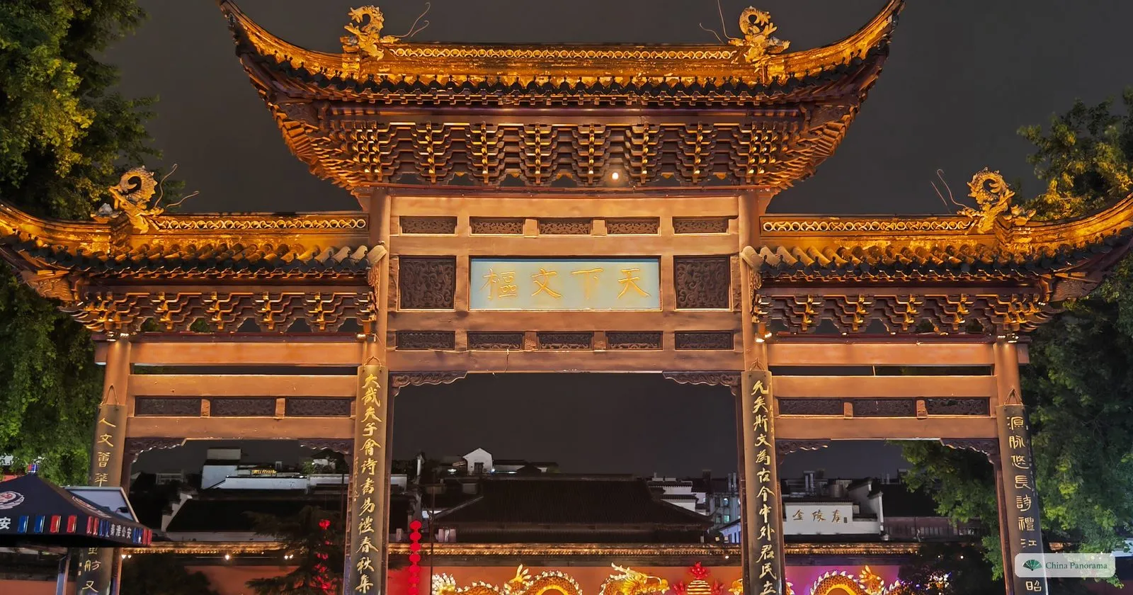 The wooden Tianxia Wenshu archway at Fuzimiao lit gold at night, dragons in coloured lights on the wall across the river