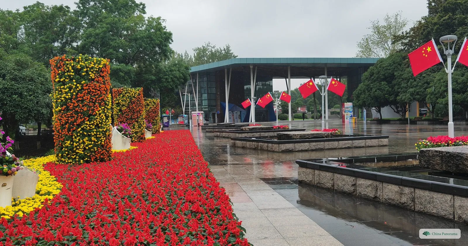 A plaza with a carpet of red flowers, columns of flowers and a row of Chinese flags, wet paving after rain