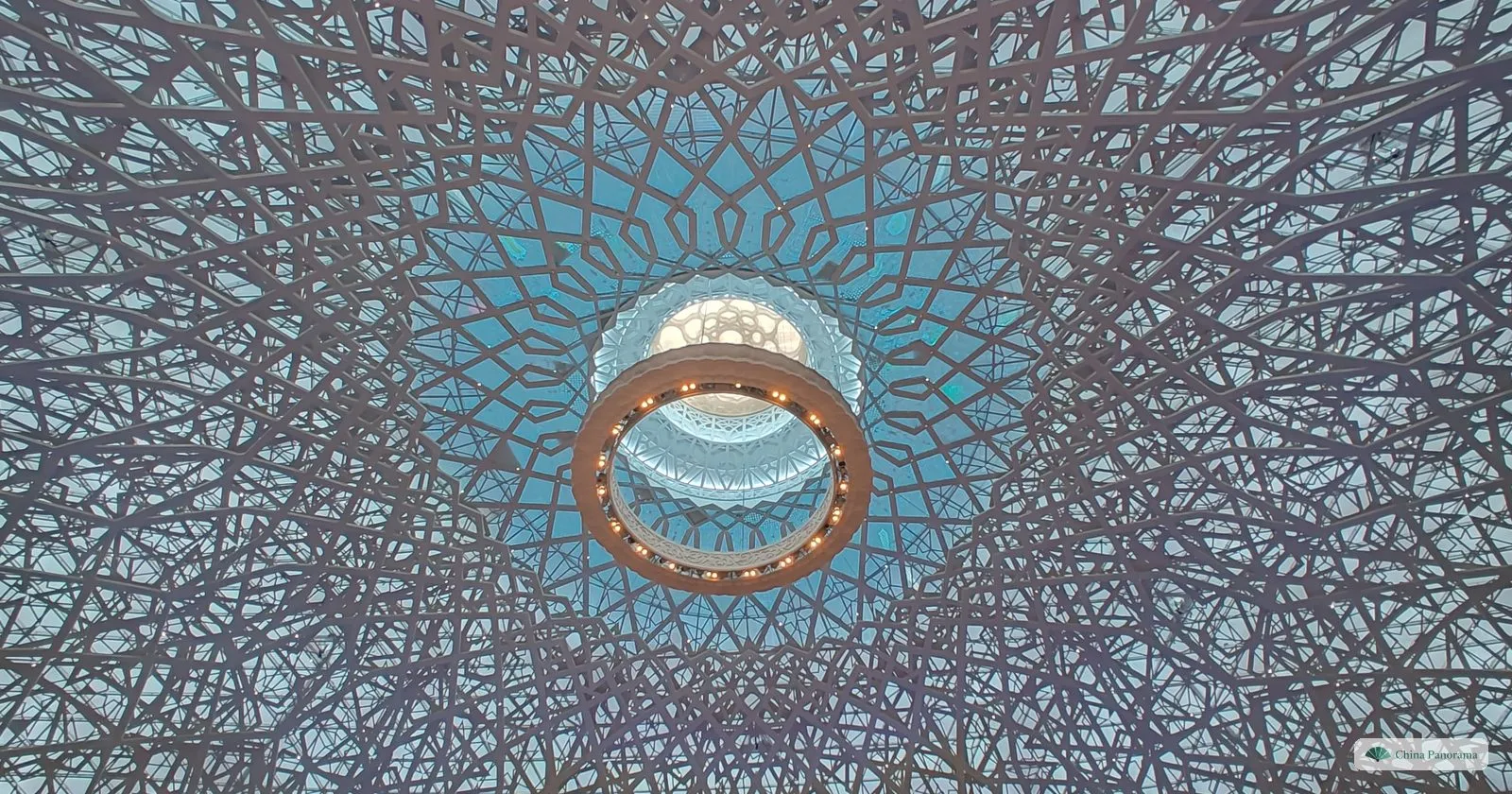 Looking straight up into a dome of interlaced steel ribs around a round skylight with a golden ring