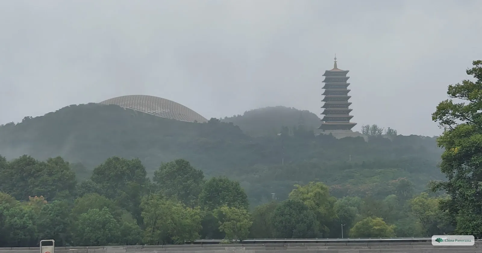 A wooded mountain in mist with a tall many-tiered pagoda on one ridge and a pale dome rising behind the trees