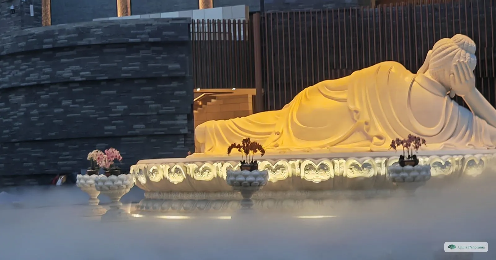 A white jade reclining Buddha lit golden on a lotus base, half hidden in a bank of artificial mist, dark stone walls behind