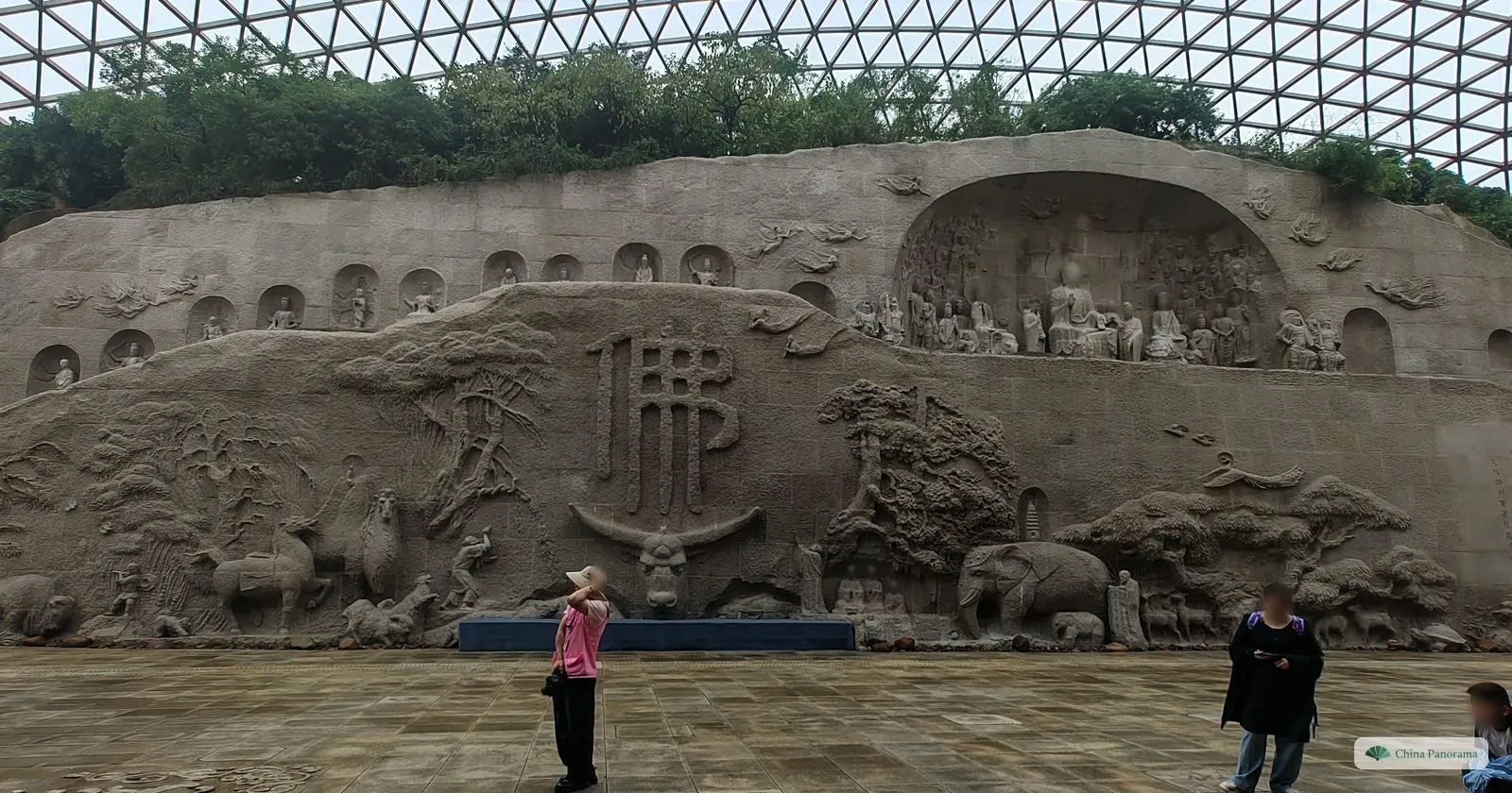 A long stone relief wall with the character 佛 above an ox head, elephants, horses and trees carved in it, niches with seated figures along the top, visitors in front