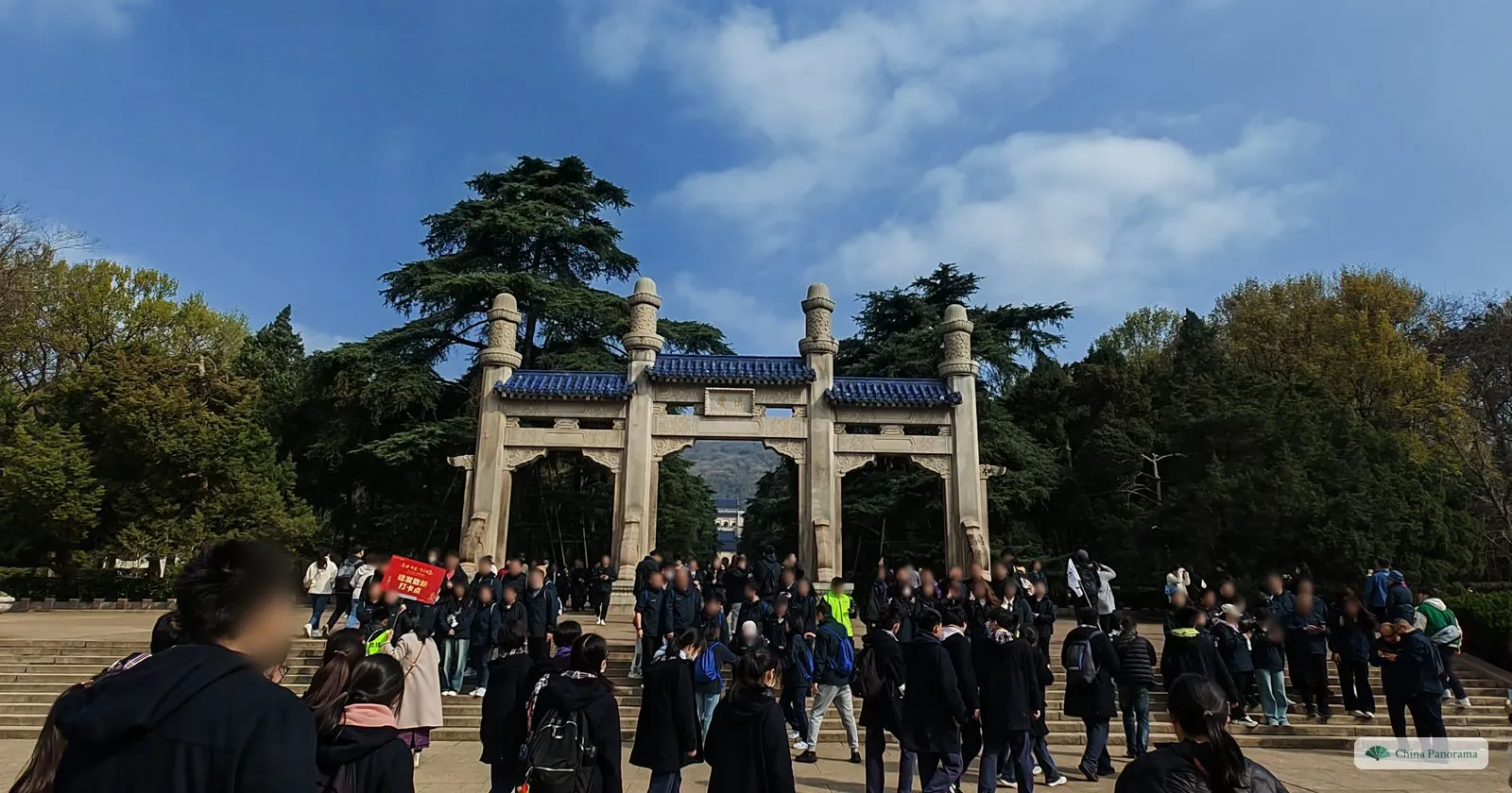 A pale stone archway with four columns and blue tiled roofs between tall cedars, a crowd of tour groups walking through it