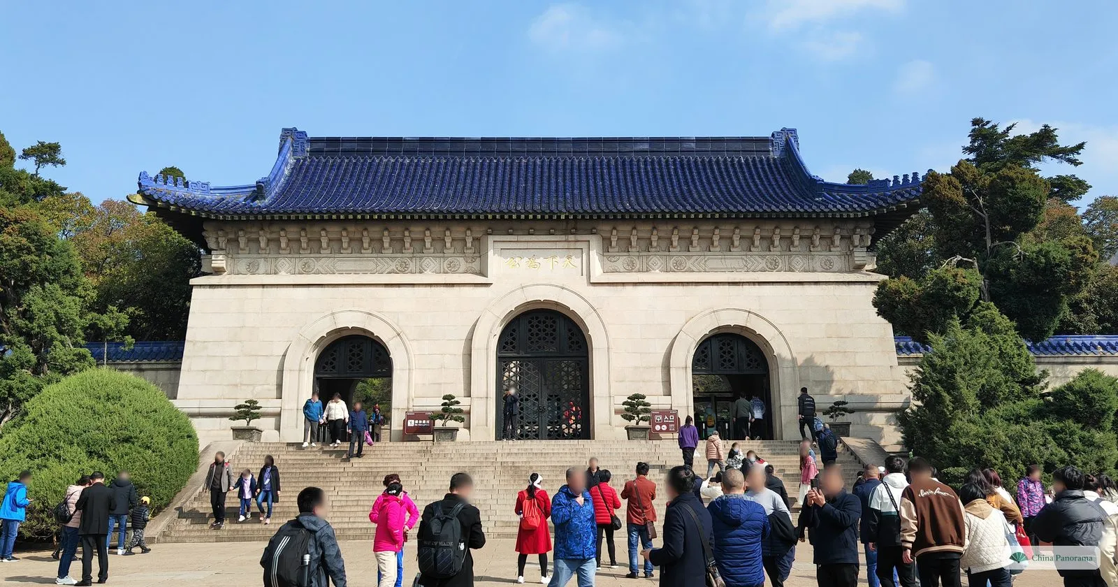 A massive pale stone gatehouse with three arched doorways and a blue glazed tile roof, gold characters above the central arch and visitors on the steps in front