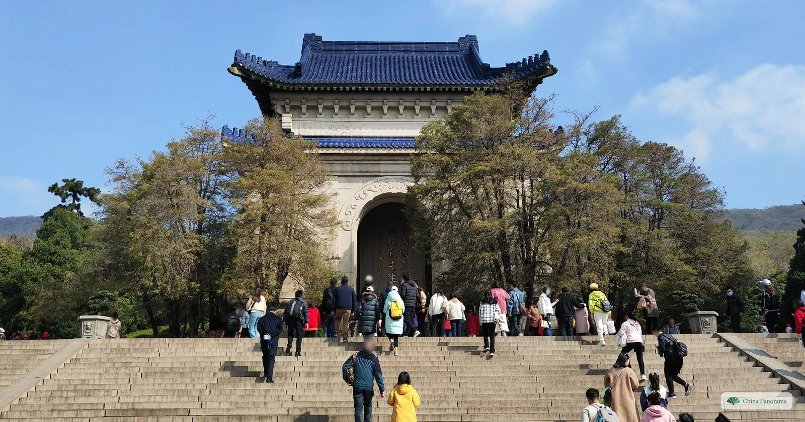 A square stone pavilion with a blue roof and a tall arched opening, at the top of wide steps with visitors climbing