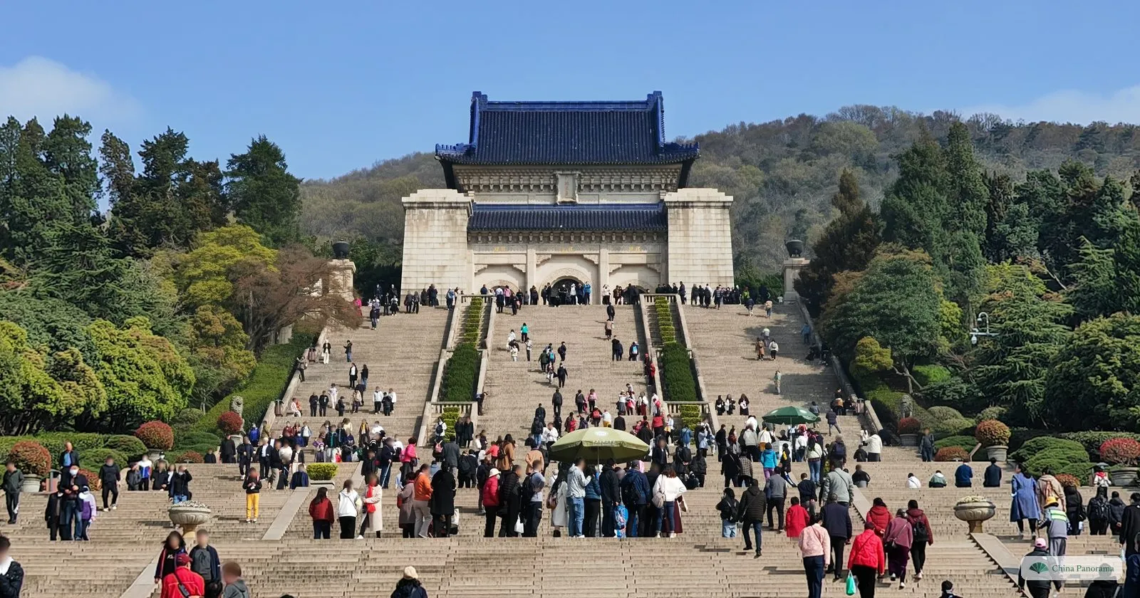The granite steps of the Sun Yat-sen Mausoleum climbing a wooded hillside to a pale hall with a blue tiled roof, crowds on the platforms