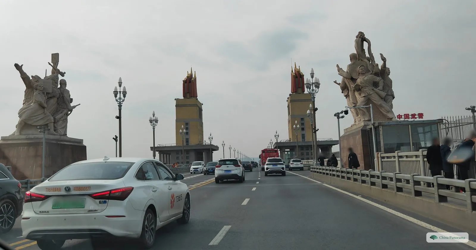 A wide road across a bridge, a large white sculpture group on a pedestal at each side, and two tall yellow towers topped with red steel flags ahead