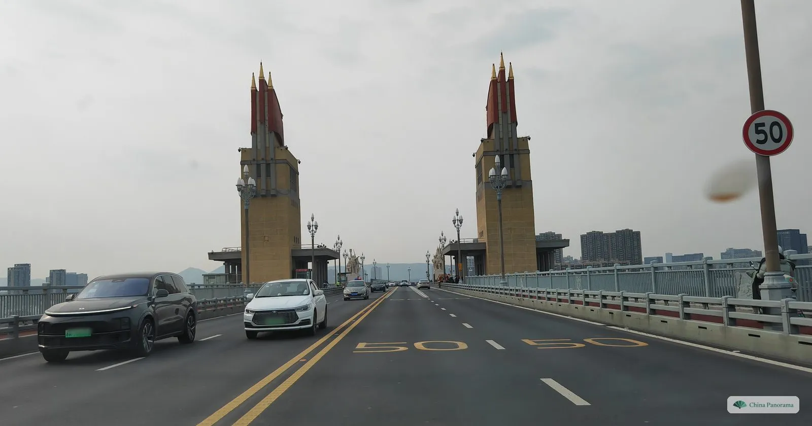 Two tall yellow towers with red steel banners on top rising either side of a long straight bridge road with cars, the number 50 painted on the asphalt