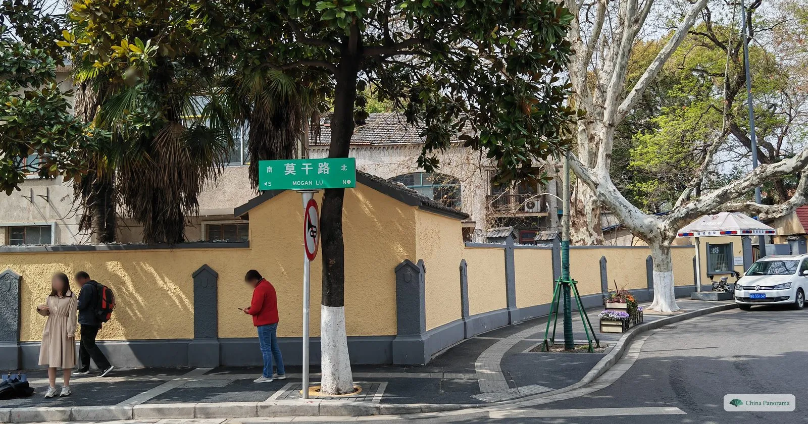 A street corner with a long yellow wall, a green street sign reading 莫干路, a palm tree and a few pedestrians