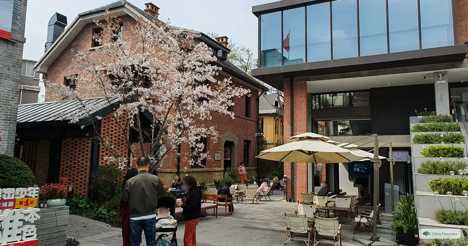 A red brick house with a white-flowering cherry tree, café tables and parasols in the courtyard and a modern glass building beside it