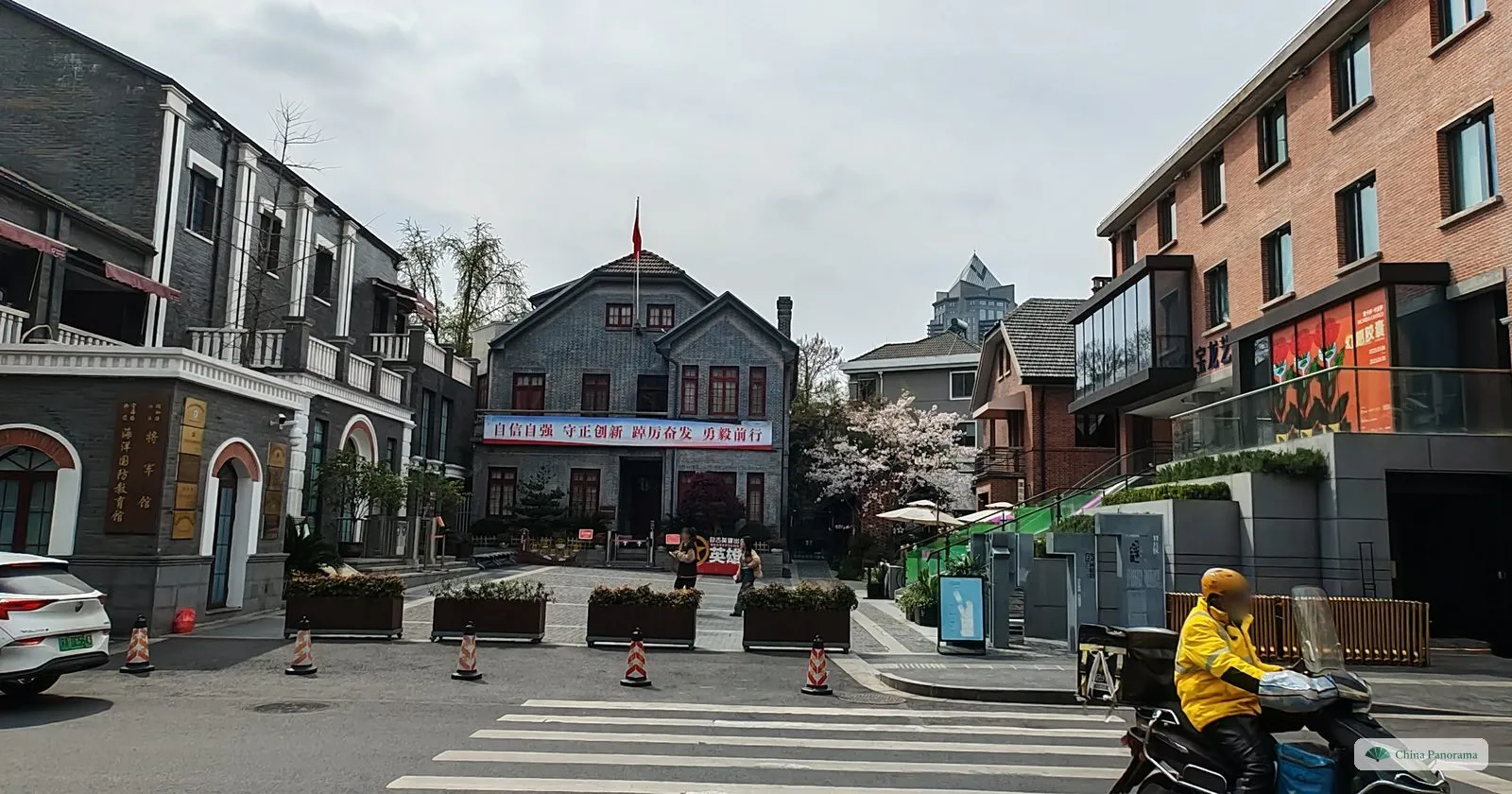 A small square surrounded by grey and red brick villas with steep roofs, a cherry tree in flower and a delivery rider passing