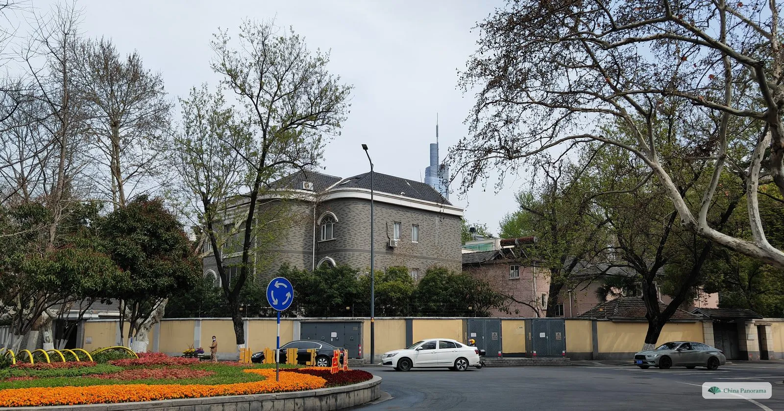 A grey stone villa with a pitched roof behind a long yellow wall, plane trees and a roundabout planted with flowers