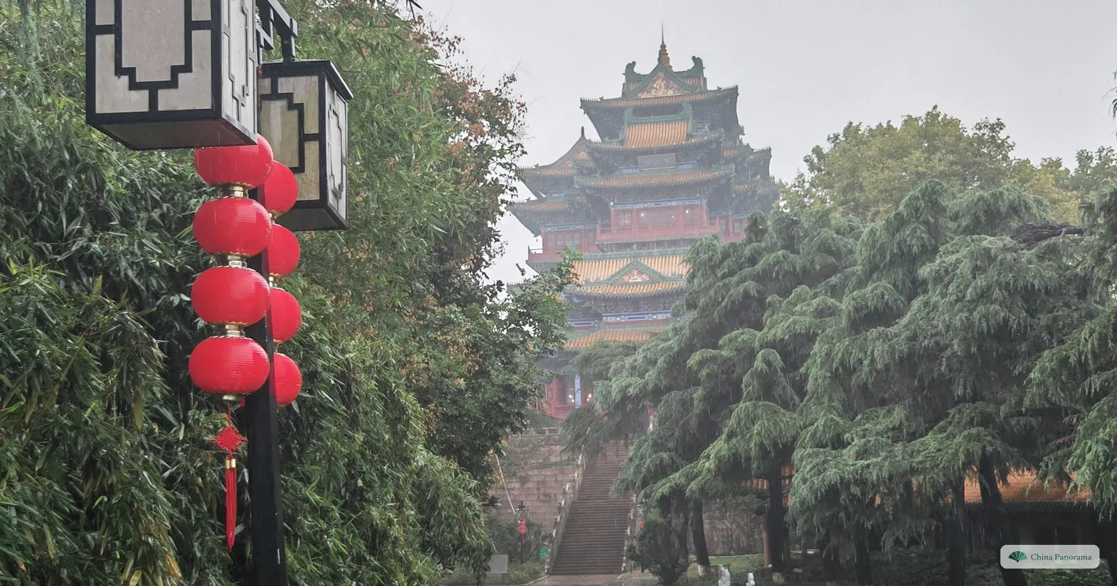 A many-tiered tower with yellow and green roofs rising out of trees and mist above a stone stair, red lanterns in the foreground