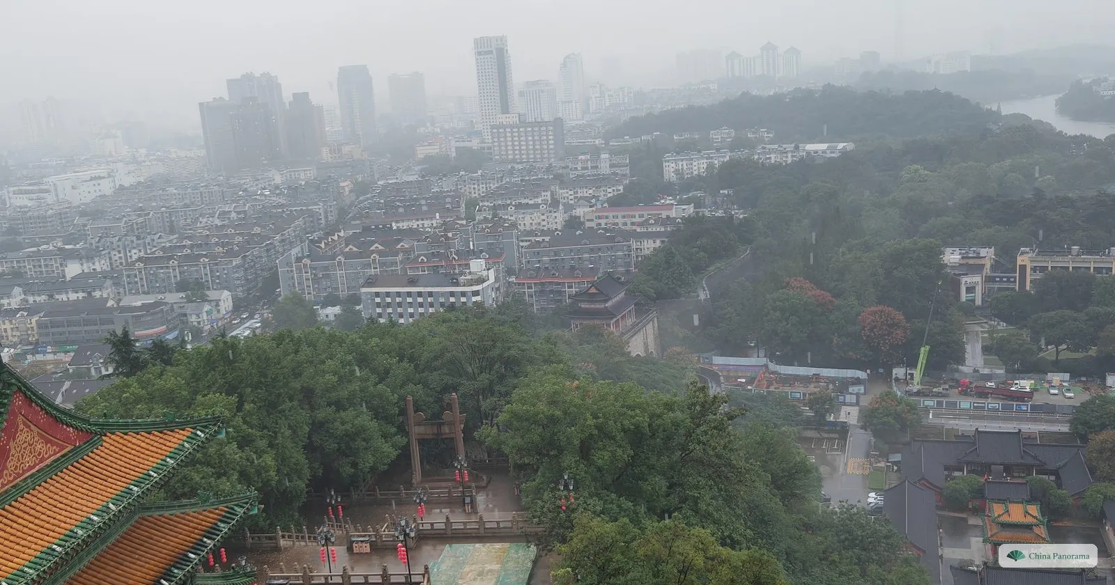 View from a high window over a hazy city of low buildings and green hills in the rain, the corner of a yellow tiled roof in the foreground