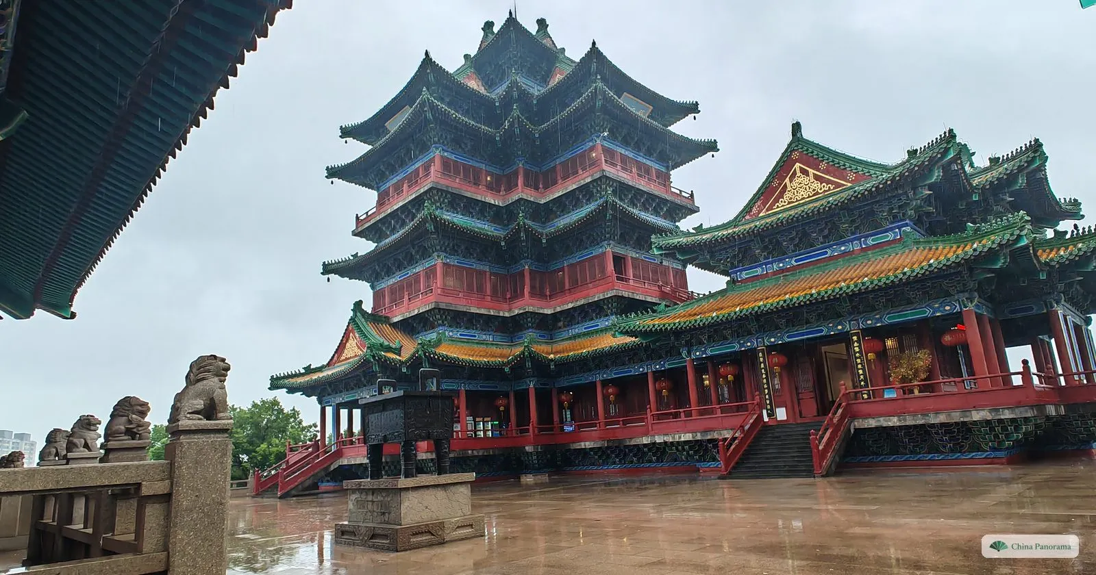 A large tower with four tiers of green-edged roofs and red pillars on a wet stone terrace, stone lions on a balustrade in the foreground