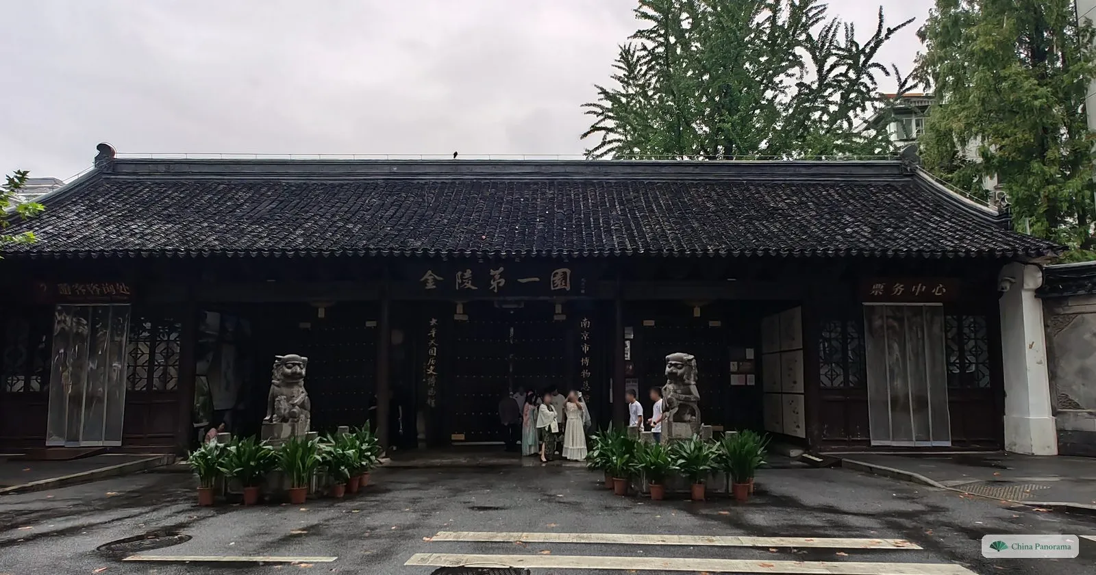 A long single-storey gatehouse with a dark tiled roof and a black plaque 金陵第一園, two stone lions and potted plants in front