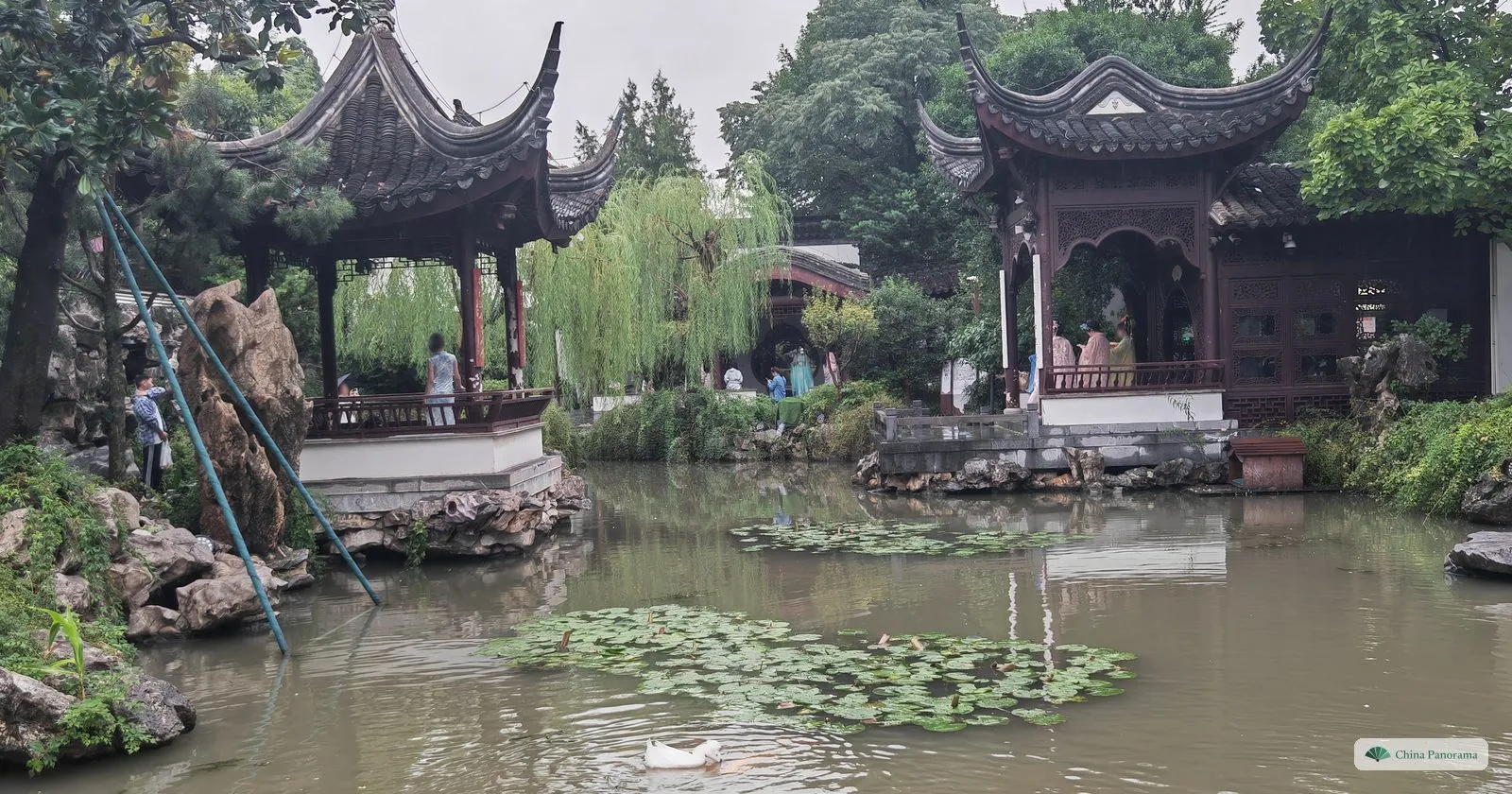 A garden pond with lily pads and two ornate pavilions with upturned eaves, rockeries and weeping willows, on a grey rainy day