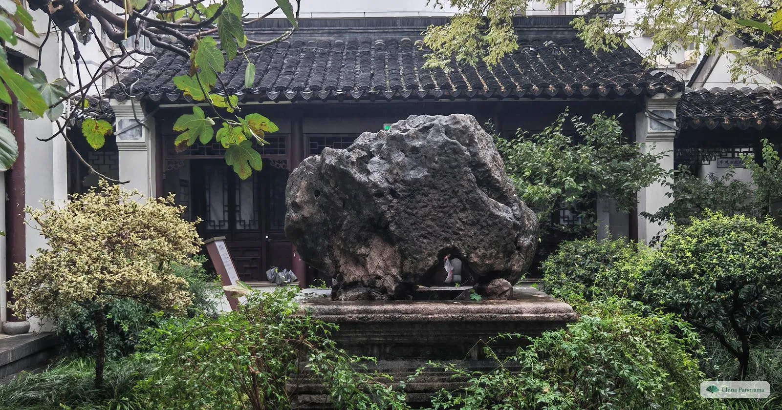 A large dark scholar’s rock on a stone base in a courtyard garden, with shrubs and a hall with wooden lattice windows behind
