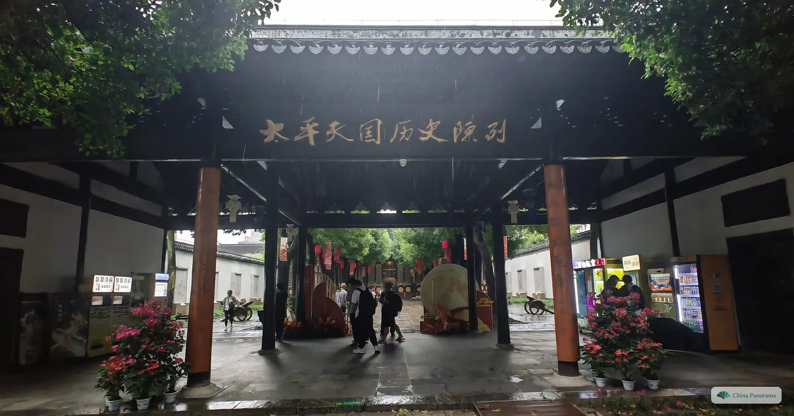 A covered gateway with a black board reading 太平天国历史陈列, looking through to a courtyard with flowers and a ticket desk