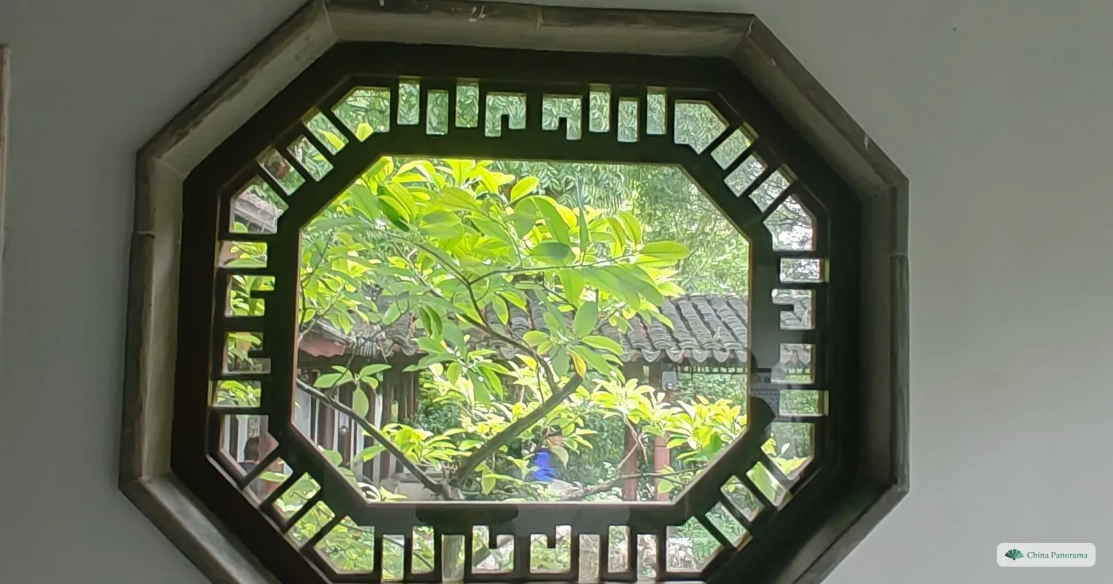 An octagonal window with a lattice frame in a white wall, green leaves and a tiled roof seen through it