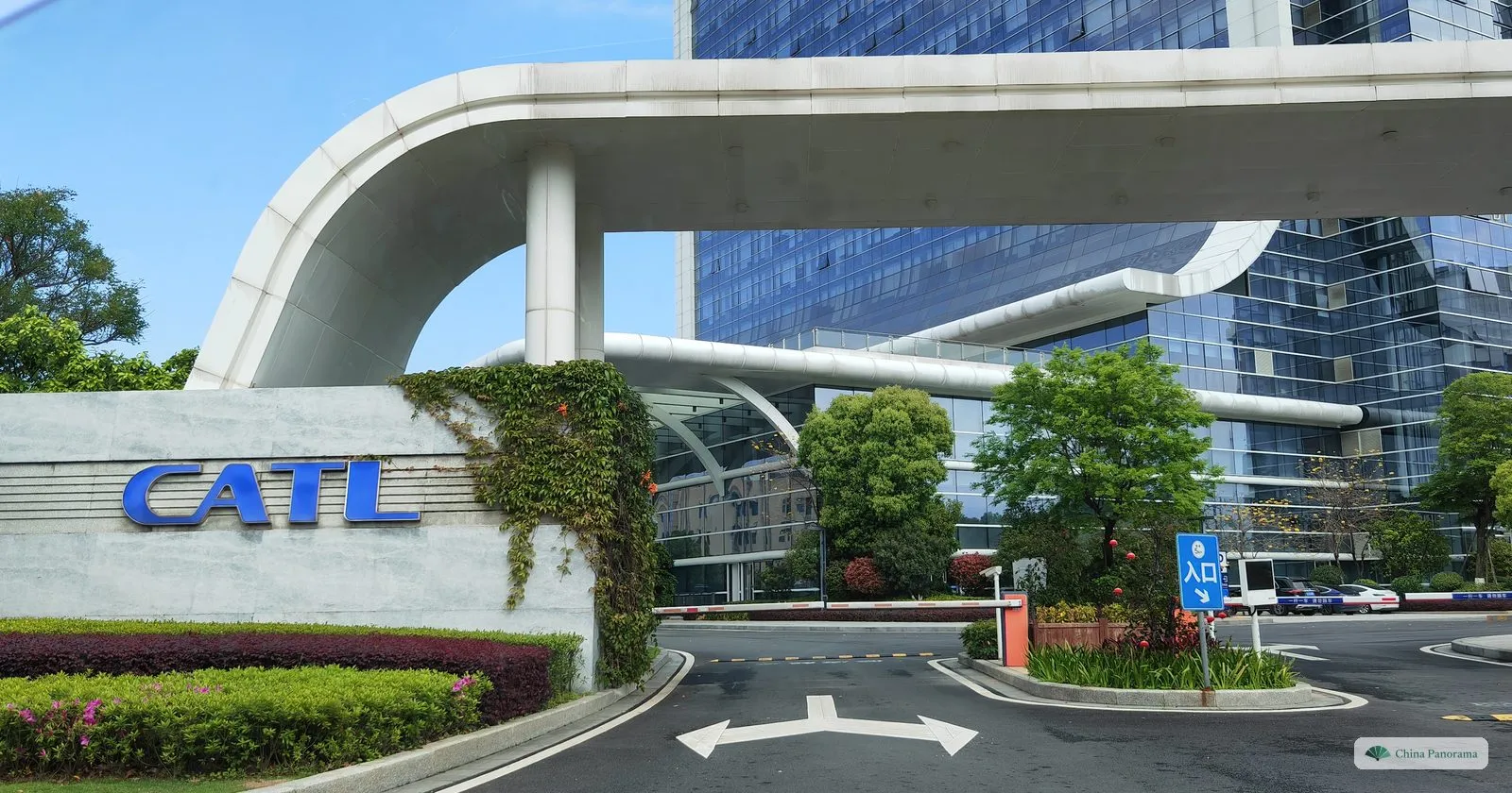 A swooping white canopy over an entrance road, a stone wall with the blue letters CATL, a barrier and an 入口 (entrance) sign, and a glass tower behind