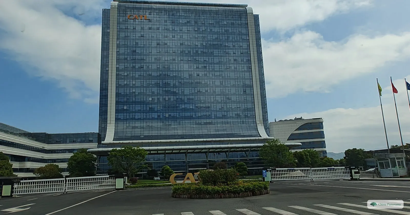 A tall glass tower with curved white edges and a small CATL logo at the top, behind a closed gate, a roundabout and three flagpoles