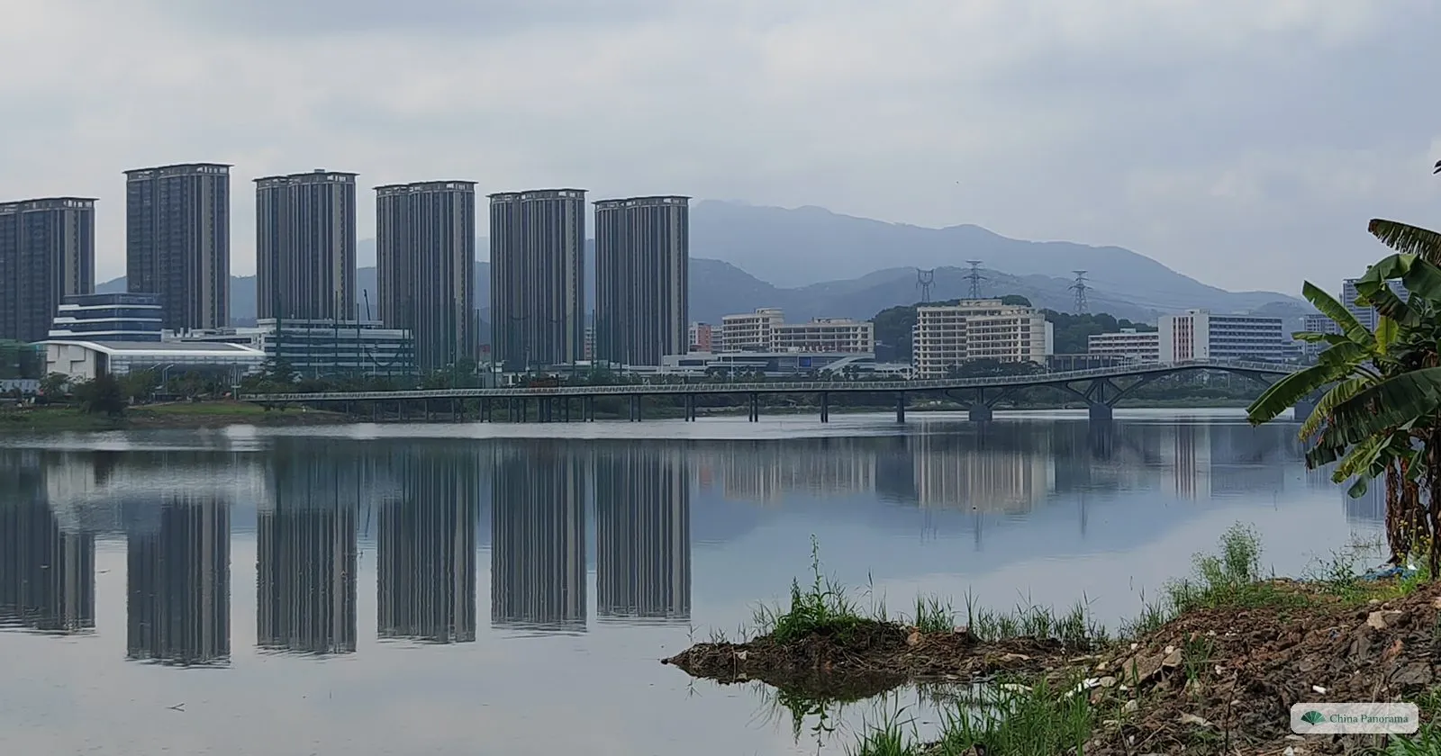 Six identical tower blocks and a road bridge reflected in a still lake, with green hills behind and a banana plant on the bank