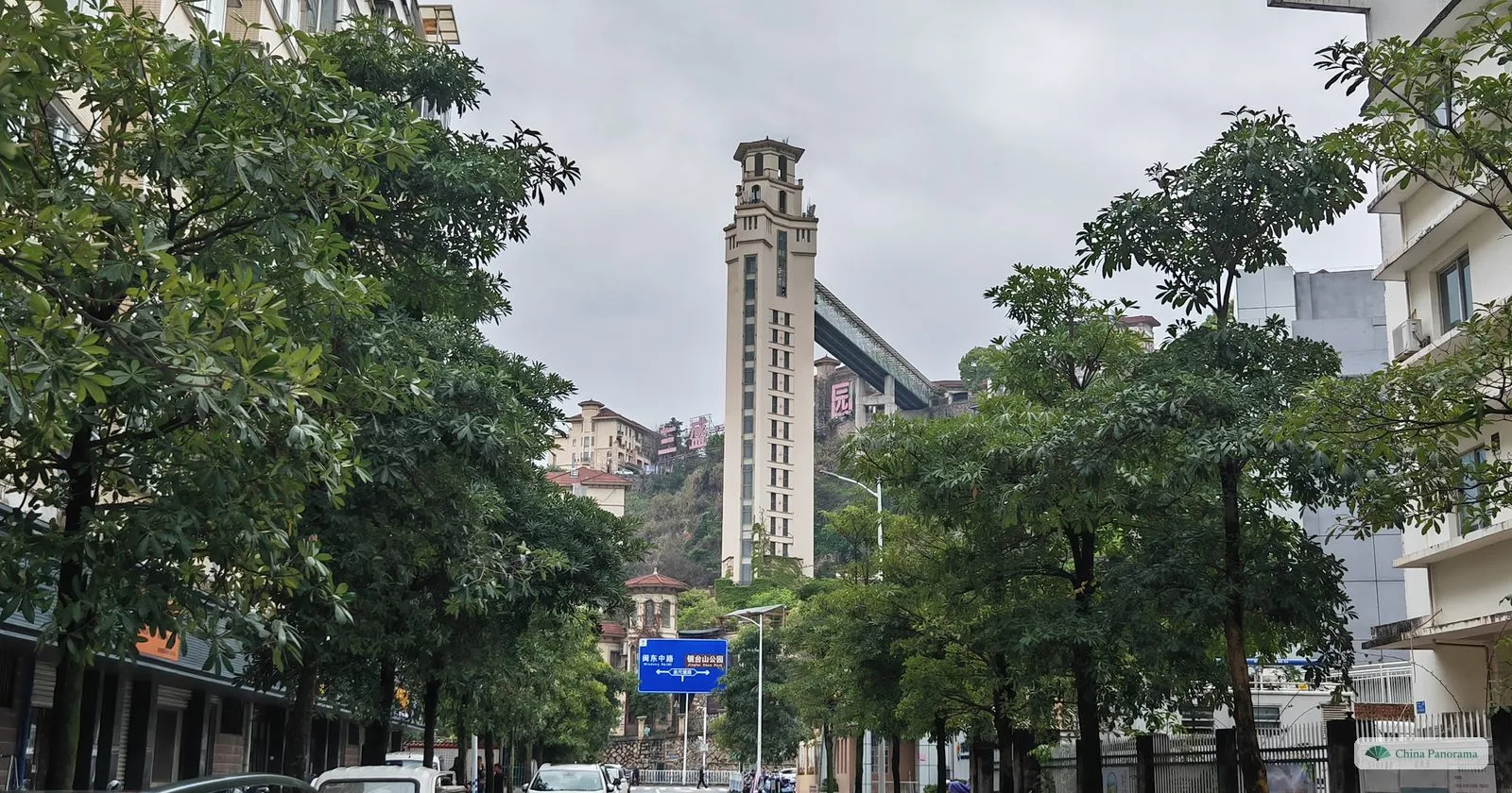 A slender cream tower about twenty storeys high joined by a long covered bridge to a green hillside, between street trees, with a blue sign in Chinese and English at its foot